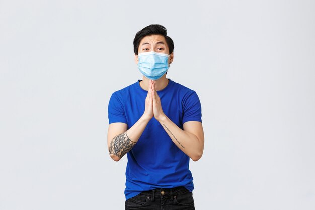 Different emotions, social distancing, self-quarantine on covid-19 and lifestyle concept. Asian guy in need desperate begging for help, hold hands in pray, pleading, wearing medical mask