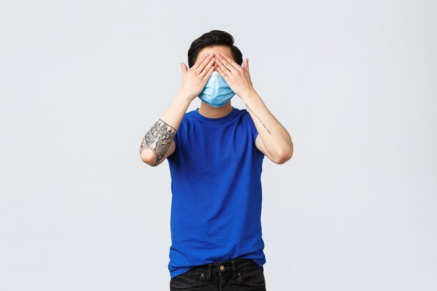 Different emotions, social distancing, self-quarantine on covid-19 and lifestyle concept. Asian man in blue t-shirt and medical mask standing blindfolded, cover eyes not see what happening