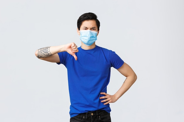 Different emotions, social distancing, self-quarantine on covid-19 and lifestyle concept. Disappointed and upset serious asian male in blue t-shirt, medical mask show thumbs-down