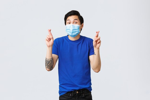 Different emotions, social distancing, self-quarantine on covid-19 and lifestyle concept. Hopeful excited asian man anticipating good news, cross finger wear medical mask