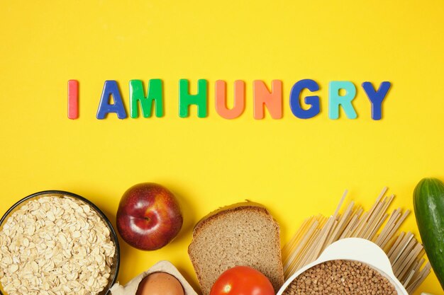 Different food items, blue measuring tape and inscription I AM HUNGRY on yellow background, eating disorder concept top view