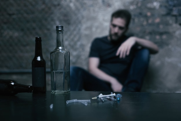 Различные полные большие бутылки, расположенные на столе в гараже рядом с использованными шприцами, пока наркоман сидит