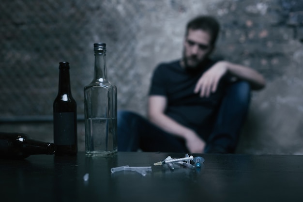 Different full big bottles located on the table in the garage next to the used syringes while junkie sitting