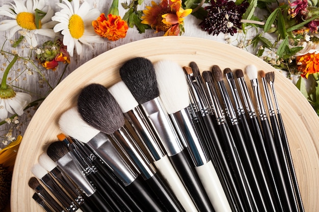 Different Make up brushes on plate next to wild flowers on wooden background