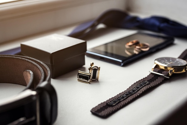 Different men's accessories such as cufflinks watches rings belt tie and phone are on the table