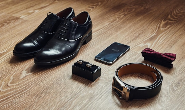 Diversi accessori da uomo come scarpe, cintura, papillon, gemelli e telefono, sono sul tavolo