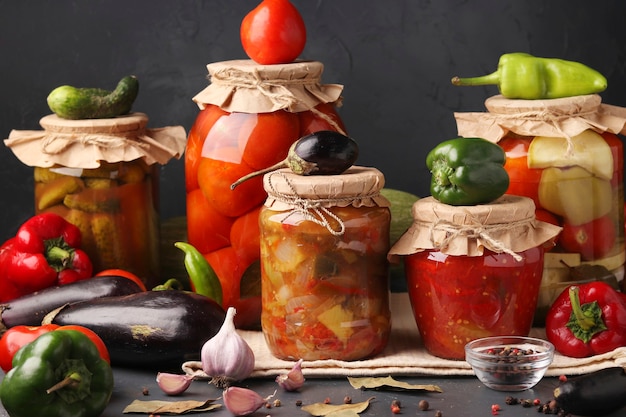 Different pickled vegetables in glass jars for long-term storage: salad with eggplant, peppers in tomato sauce, cucumbers, tomatoes and mixed vegetables against a dark surface, horizontal format