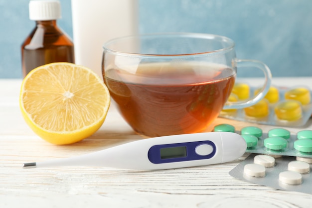 Different pills, thermometer, medical bottles, tea and lemon on white wooden table, closeup