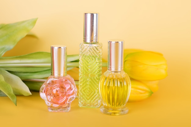 Different transparent perfume bottles with bouquet of tulips on yellow background. Aromatic essence bottles