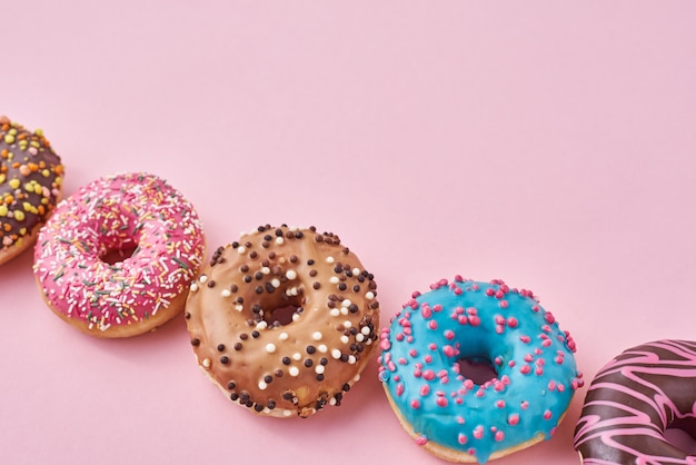 Different types of colorful donats decorated sprinkles and icing on pastel pink surface