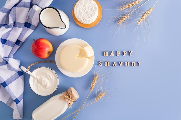 Photo different types of dairy products in bowls bottles and jugs on a blue background top view flat lay wooden letters with the text shavuot