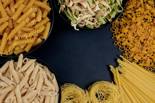 Different types of dry pasta on the plate and in bowls on black background. Space for text, top view.