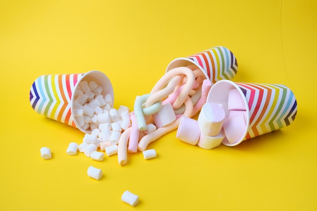 Diversi tipi di marshmallow caduti da bicchieri di carta festivi con motivo geometrico su sfondo giallo, spazio di copia