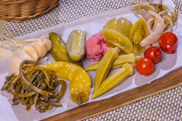 Different types of pickled vegetables on a white plate. Pickled cucumbers, tomato cherry, greens, mushrooms and garlic with onion and lettuce on the table.