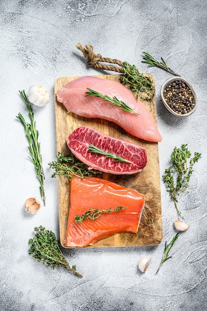 Different types of raw meat on cutting board with herbs