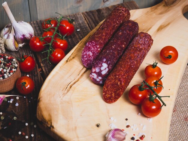 Different types of salami on a wooden board with spices and\
cherry tomatoes