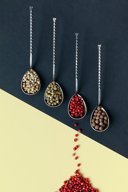 Different varieties of peppercorns in metal spoons on a double-colored paper surface