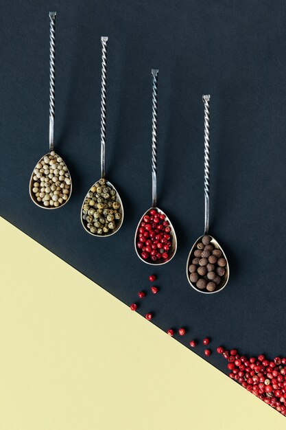 Different varieties of peppercorns in metal spoons on a double-colored paper surface