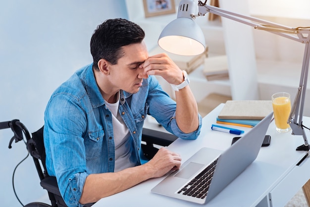 Difficult task. Thoughtful invalid man keeping eyes closed and putting arms on table while sitting at his workplace