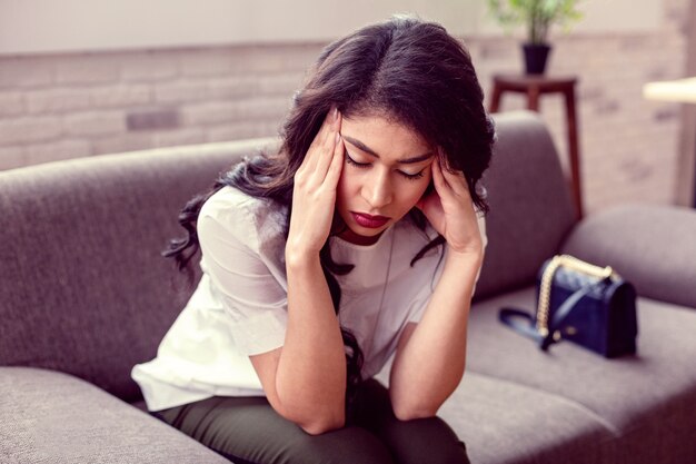 Difficult times. Pleasant brunette woman touching her temples while sitting at home on the sofa