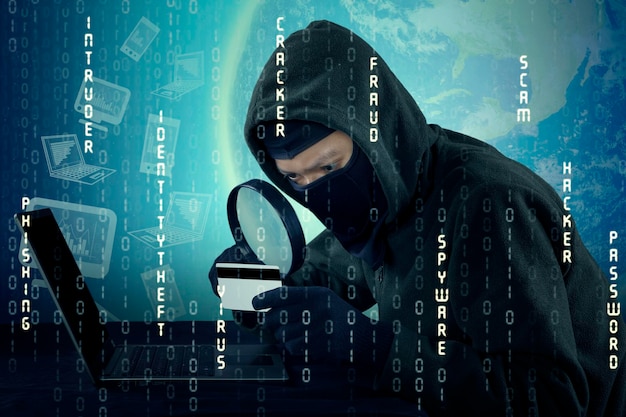 Digital composite image of computer hacker looking at card with magnifying glass while using laptop