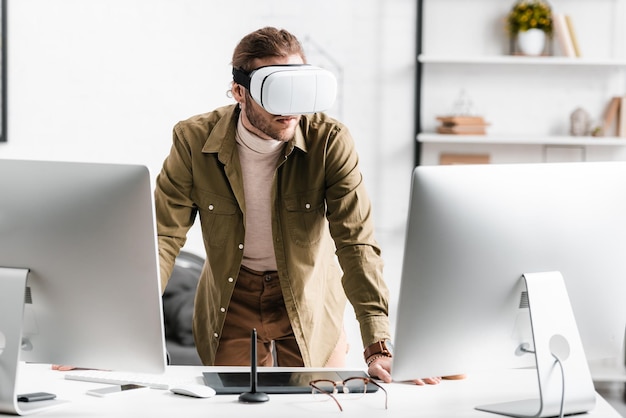 Digital designer in virtual reality headset standing near computers and graphics tablet on table