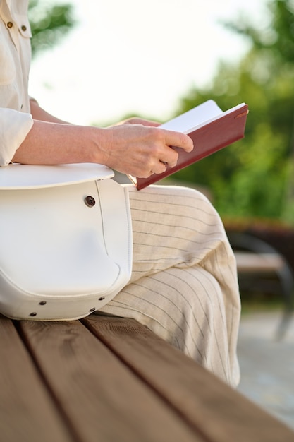 Digital detox. Woman in light trousers reading notes sitting on park bench on fine day, no face