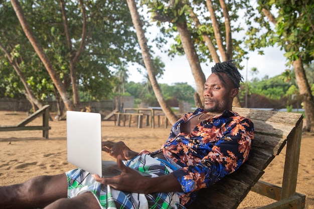 A digital nomad consults his computer while lying on the bench by the sea