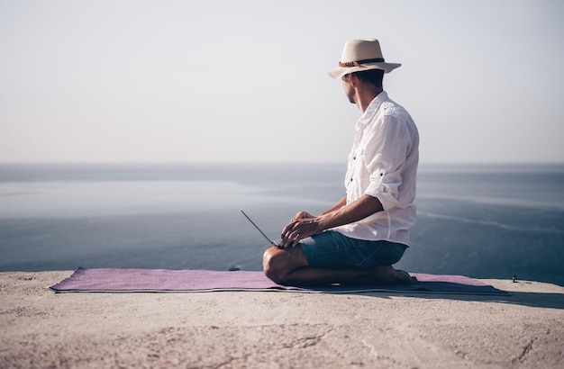 A digital nomad a man in a hat a businessman with a laptop does yoga on the rocks by the sea at