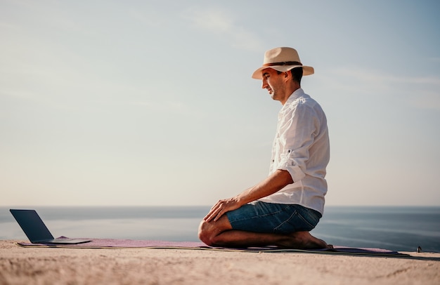 A digital nomad a man in a hat a businessman with a laptop does yoga on the rocks by the sea at