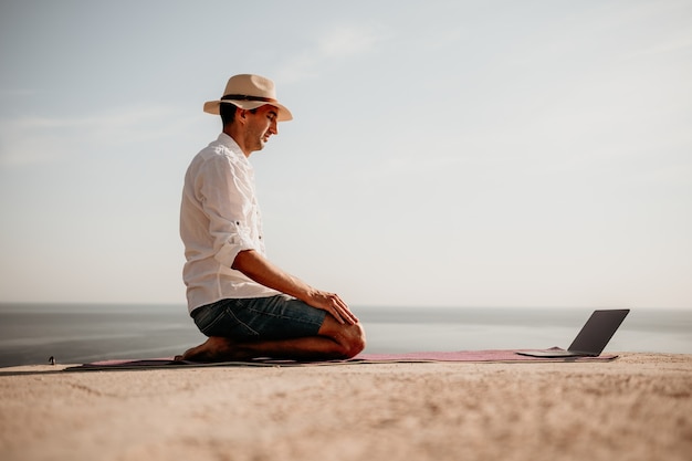 A digital nomad a man in a hat a businessman with a laptop does yoga on the rocks by the sea at