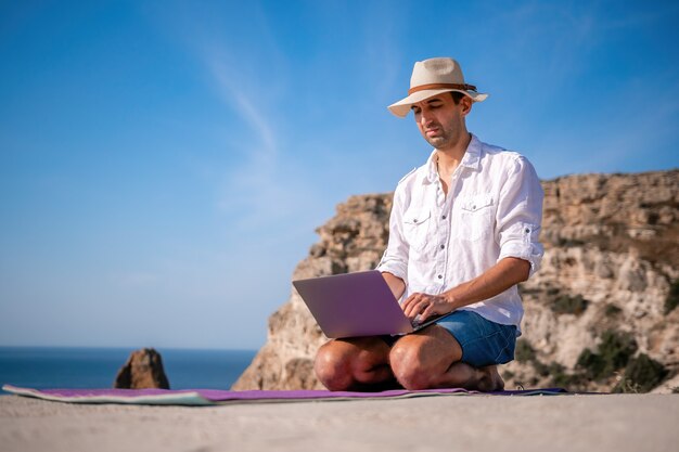 A digital nomad a man in a hat a businessman with a laptop does yoga on the rocks by the sea at