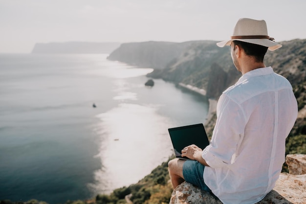 Digital nomad man in the hat a businessman with a laptop sits on the rocks by the sea during sunset