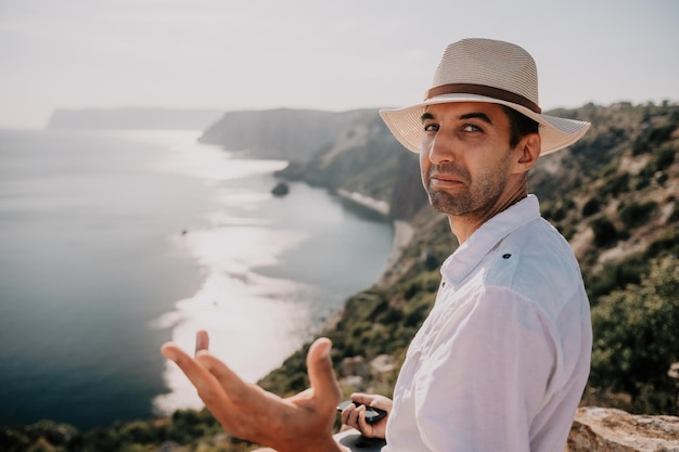 帽子をかぶったデジタル遊牧民の男ノートパソコンを持っているビジネスマンは日没時に海のそばの岩の上に座っています