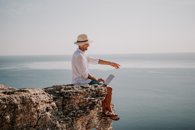 帽子をかぶったデジタル遊牧民の男ノートパソコンを持っているビジネスマンは日没時に海のそばの岩の上に座っています