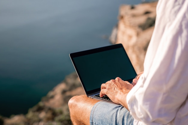 Digital nomad man in the hat a businessman with a laptop sits on the rocks by the sea during sunset