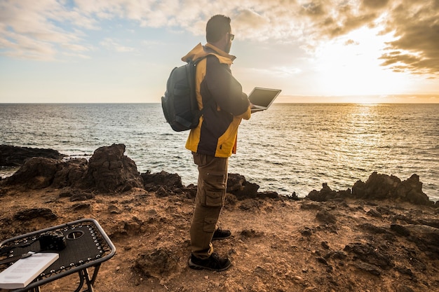 Digital nomad traveler man at work lokking an amazing golden sunset on the ocean