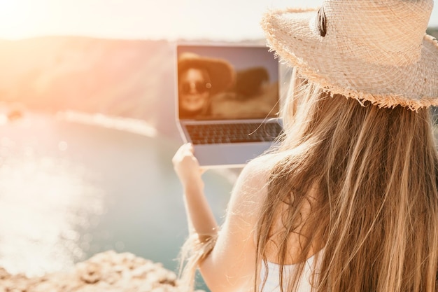 Digital nomad woman in the hat a business woman with a laptop\
sits on the rocks by the sea during