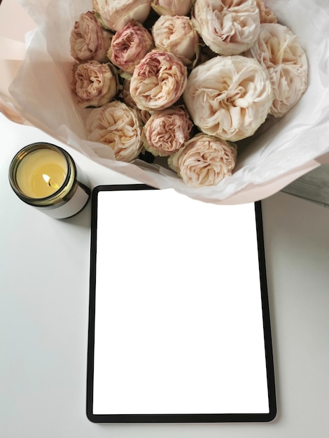 Digital tablet with a blank screen on a table with a bouquet of spray roses and a candle