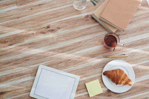 Digital tablet with empty screen, tea with pastry and stack of books on table of creative college student, view from above