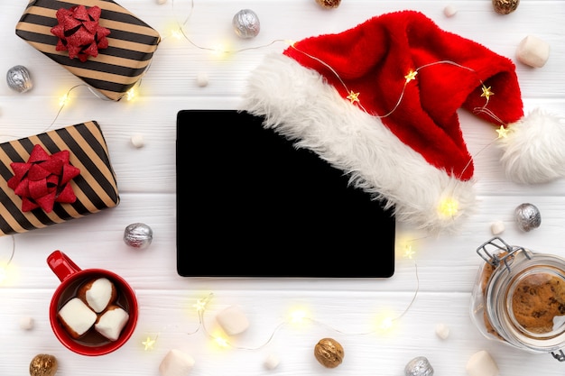 Digital tablet with hot chocolate and gifts on white wood. Christmas flat lay