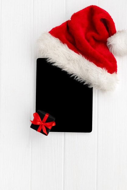 Digital tablet with Santa hat and gift on white wood. Christmas flat lay