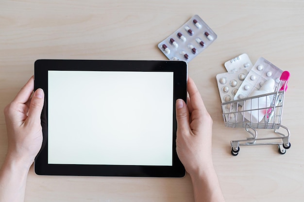 Digital tablet with a white screen in female hands and a mini cart with blisters of pills The concept of a mobile application for the remote purchase of medicines Online pharmacy