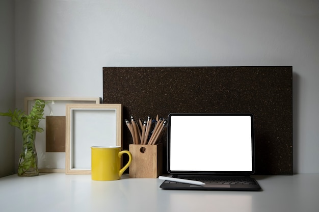 Цифровой планшет с беспроводной клавиатурой, кофейной чашкой, держателем для карандашей, комнатным растением и рамкой для рисунка на белом столе.
