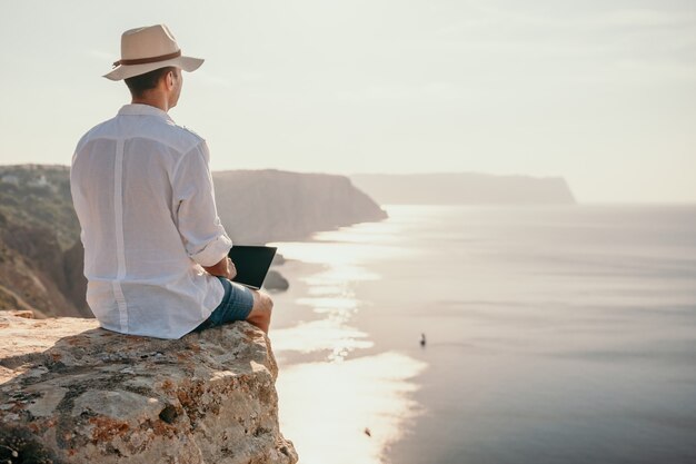 Digitale nomade, man in de hoed, een zakenman met een laptop zit op de rotsen bij de zee tijdens zonsondergang, maakt een zakelijke transactie online vanaf een afstand. Werken op afstand op vakantie.