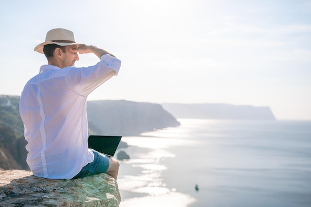 Digitale nomade man in de hoed een zakenman met een laptop zit op de rotsen bij de zee tijdens zonsondergang