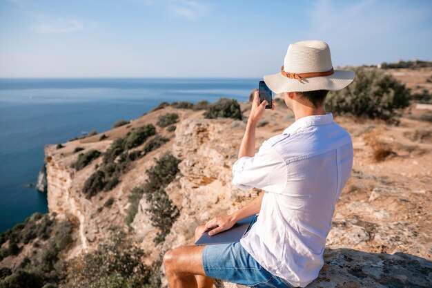 Digitale nomade man in de hoed een zakenman met een laptop zit op de rotsen bij de zee tijdens zonsondergang