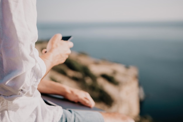 Digitale nomade man in de hoed een zakenman met een laptop zit op de rotsen bij de zee tijdens zonsondergang