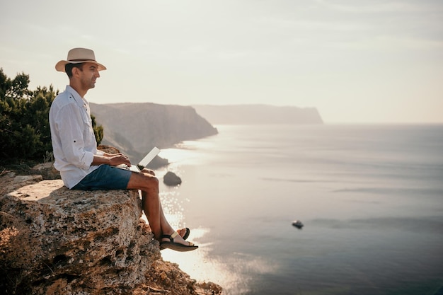 Digitale nomade man in de hoed een zakenman met een laptop zit op de rotsen bij de zee tijdens zonsondergang