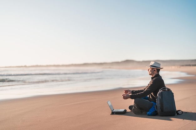 Digitale nomade zittend op het strand mediteren met zijn laptop aan de kust van het strand bij zonsondergang